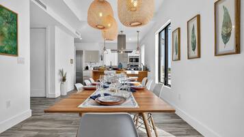 Uma área de refeições moderna com uma mesa de madeira posta para seis pessoas, paredes brancas e uma cozinha ao fundo.