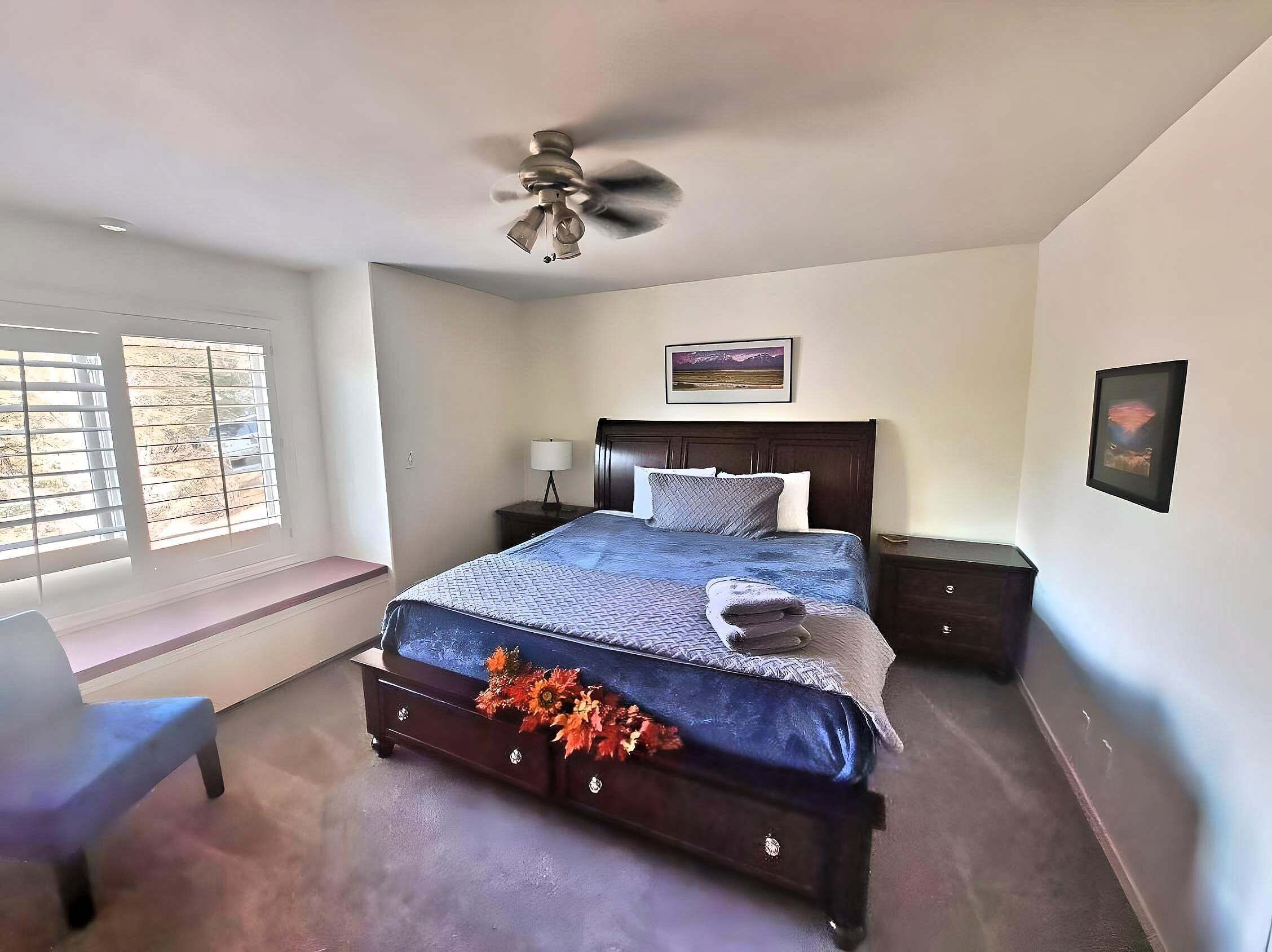 Un dormitorio con cama, ventilador de techo, mesita de noche, silla y ventana con persianas.