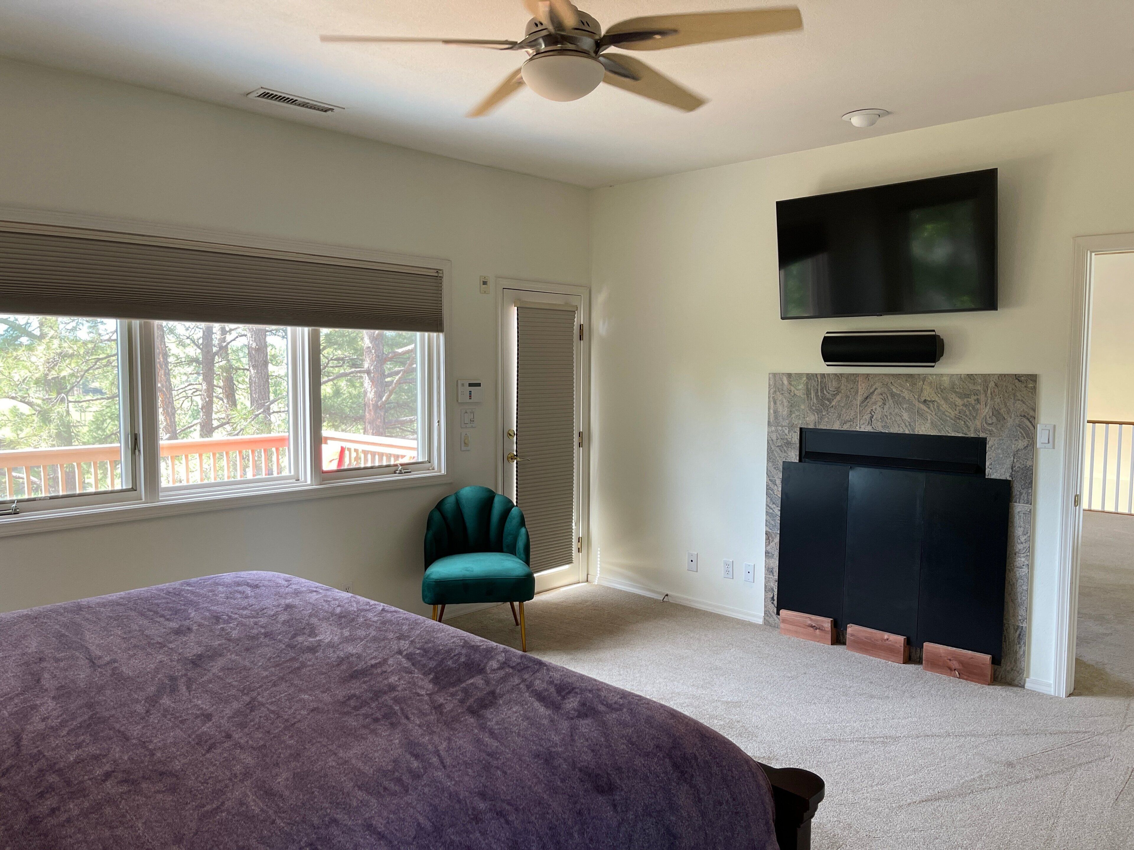 A bedroom with a bed, a TV mounted on the wall, a ceiling fan, and a window with blinds.
