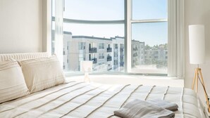 Ein Schlafzimmer mit großem Fenster, einem Bett mit weißer Bettwäsche und einer Nachttischlampe.