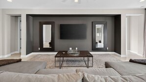 Un salon moderne avec un téléviseur à écran plat fixé au mur, un mur d’accent de couleur foncée, une table basse en bois et un espace salon avec des canapés.