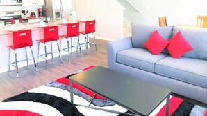 Een moderne woonkamer met een grijze bank, rode kussens, een zwarte salontafel en een keukenhoek met rode barstoelen.