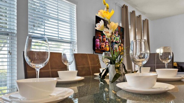 A dining table set with white plates and glasses, a vase with yellow flowers, and a painting of animated characters. - Gdl3206 (Kissimmee)