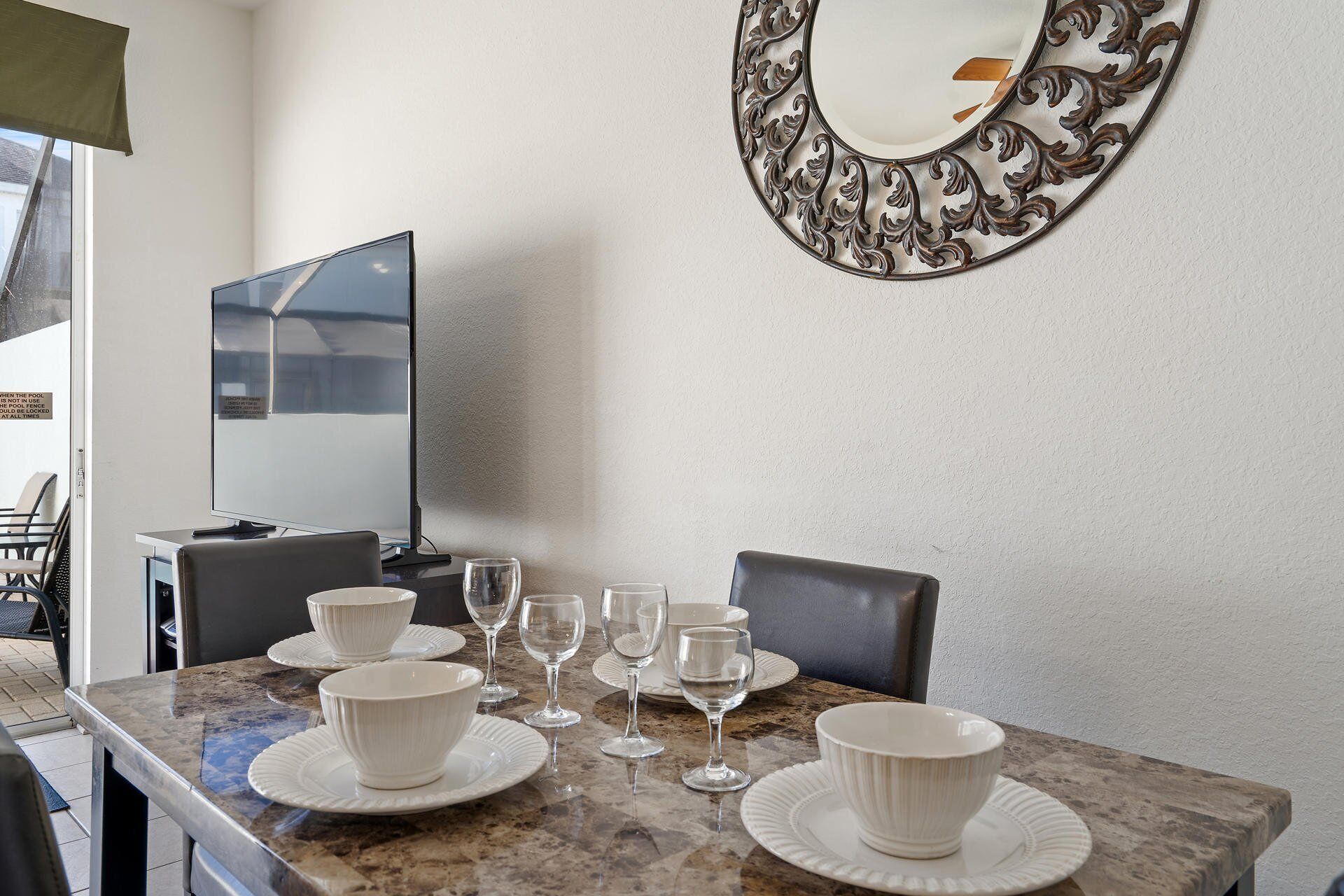 A dining area with a marble table, white plates, and wine glasses. There is a large mirror on the wall and a television on the left.