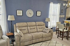 Ein Wohnzimmer mit einem beigefarbenen Sofa, zwei Nachttischen mit Lampen, einem Essbereich mit Tisch und Stühlen sowie einer Wanduhr.