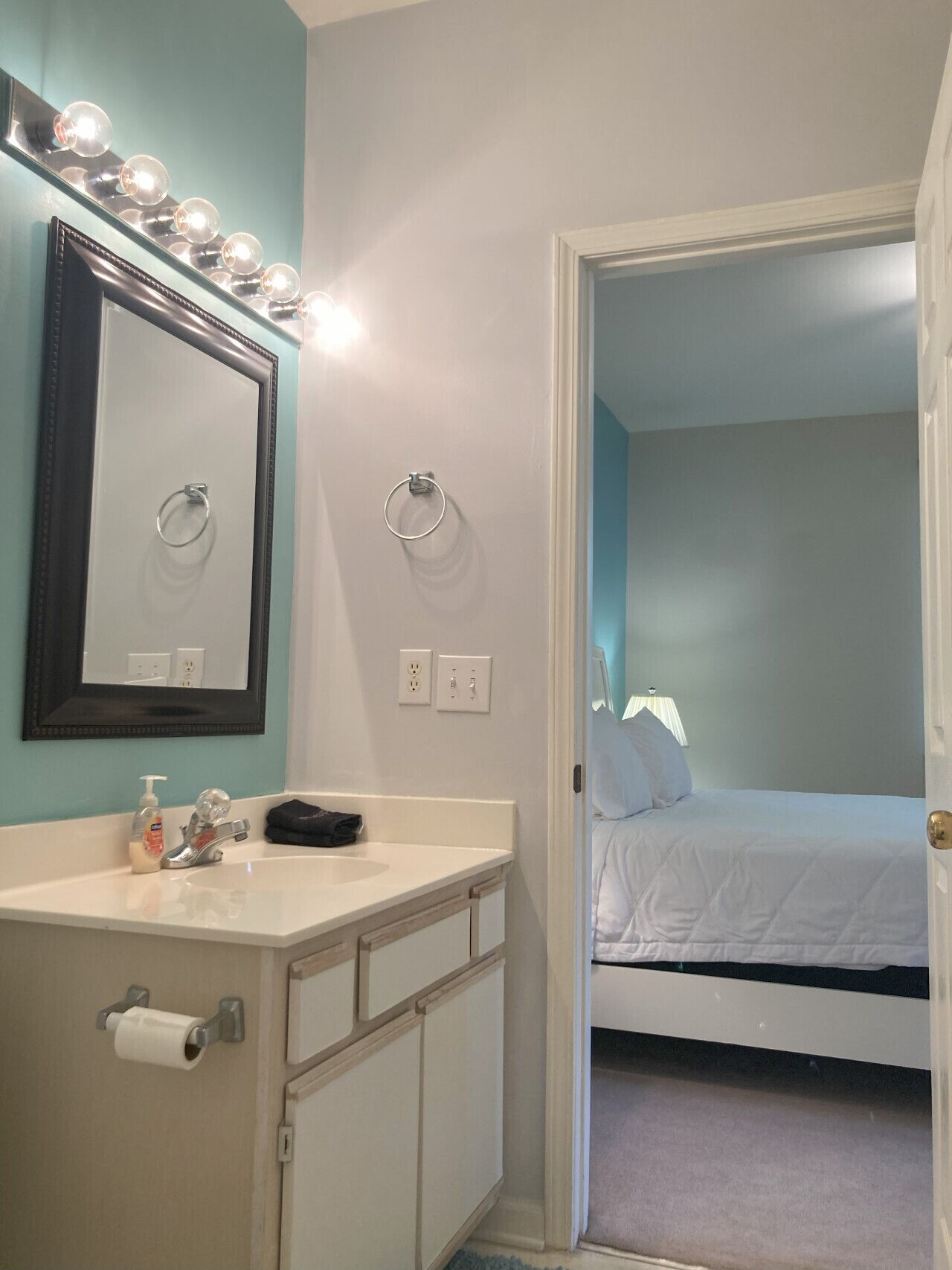 A bathroom with a vanity, mirror, and lights. There is a door leading to a bedroom with a bed and a lamp.