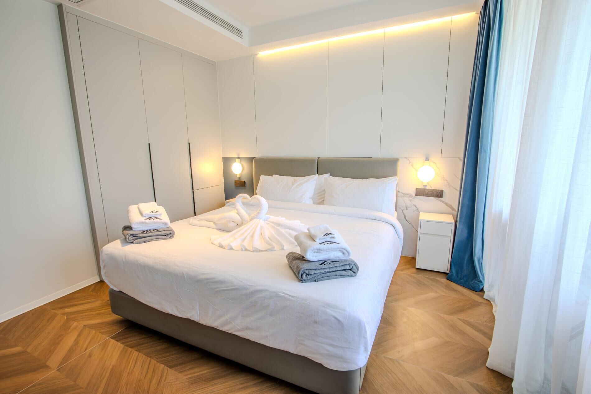 A neatly made bed with white linens and towels, a headboard, bedside tables with lamps, and a herringbone-patterned wooden floor.
