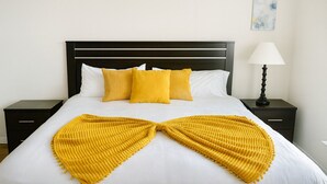Ein ordentlich bezogenes Bett mit weißer Daunendecke und zwei gelben Kissen, ein schwarzer Nachttisch mit Lampe sowie ein kleines Bild an der Wand.