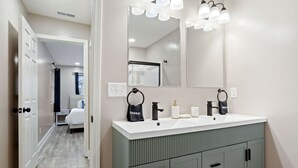 Une salle de bain moderne avec un lavabo blanc, deux robinets noirs et un grand miroir. On peut voir deux sacs noirs sur le lavabo, l’un portant l’étiquette « Maquillage » et l’autre « Manucure ». La salle de bain est équipée d’un meuble gris clair avec un plan de travail blanc.