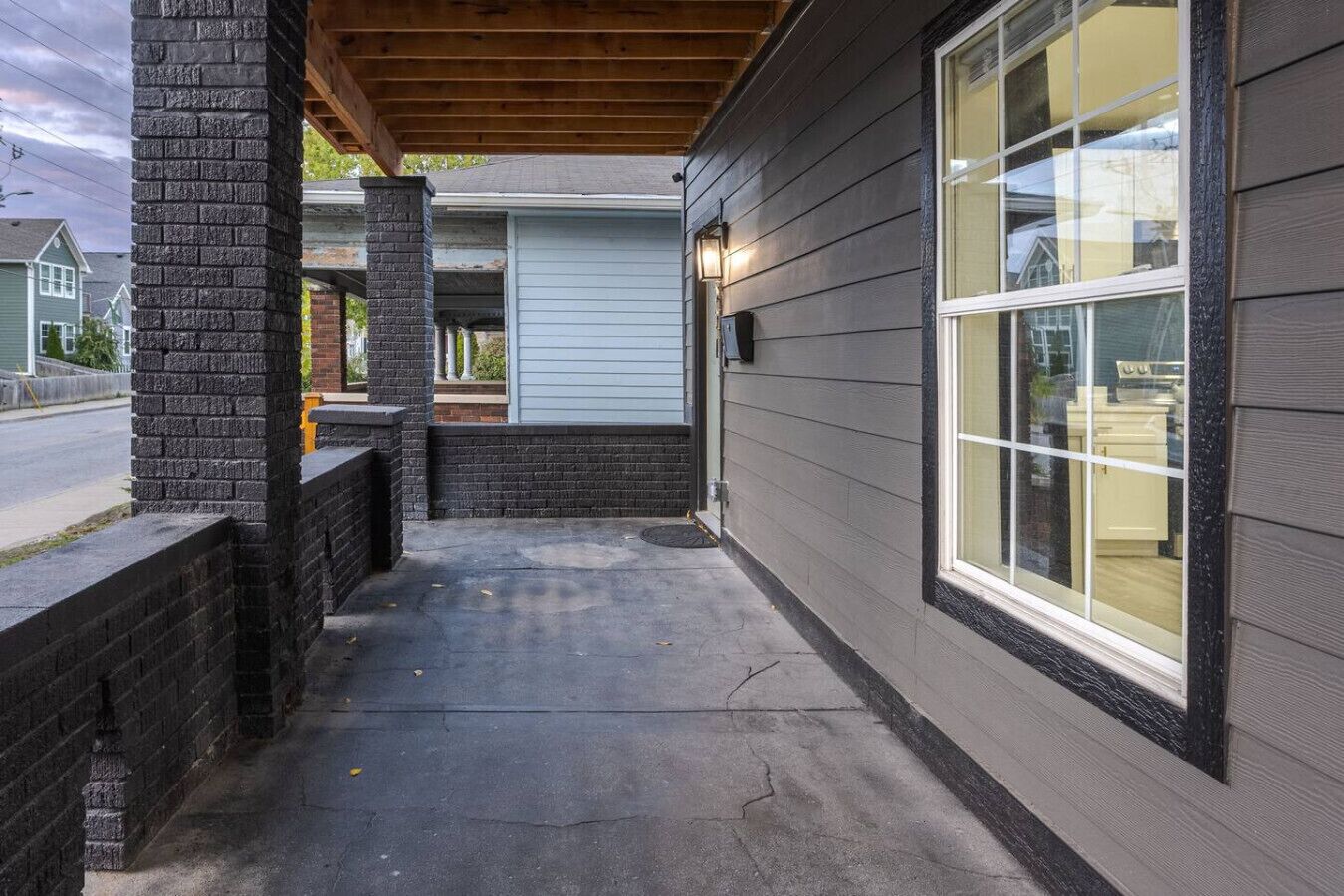 A covered porch with a brick pillar, a concrete floor, and a window with a view of a house in the background.