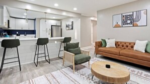 Une cuisine moderne avec des meubles blancs, un îlot central et des tabourets de bar. Le salon est agrémenté d’un canapé en cuir marron avec des coussins verts.