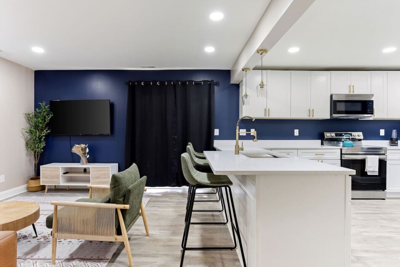 A modern kitchen with white cabinets, a black microwave, and a stainless steel stove. There is a bar area with stools and a flat-screen TV mounted on the wall.
