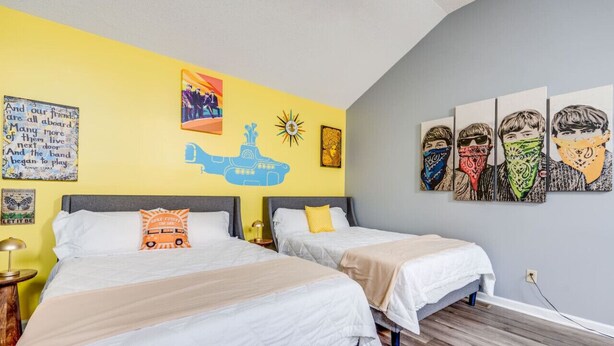 Ein Zimmer mit zwei Betten, einer gelben Wand mit Kunstwerken und einer grauen Wand mit Band-Motiven.