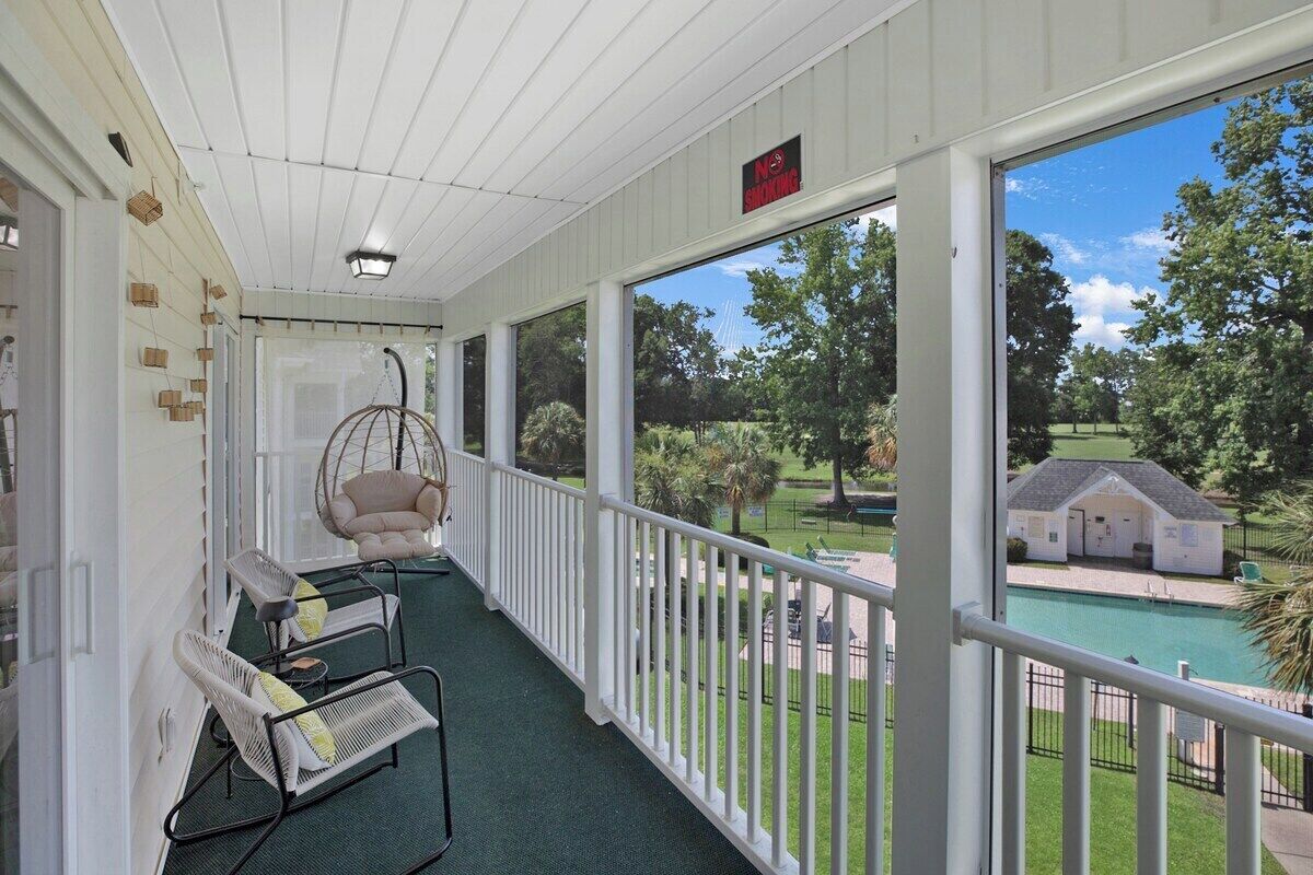 A covered porch with white railings, wicker chairs, and a hanging chair overlooking a pool and greenery.