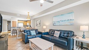 A living room with a blue leather sofa, a wooden coffee table, and a TV on a cabinet. There is a kitchen in the background with wooden cabinets and a dining area. - Sea Dunes 502 - Beach Oasis (Fort Walton Beach)
