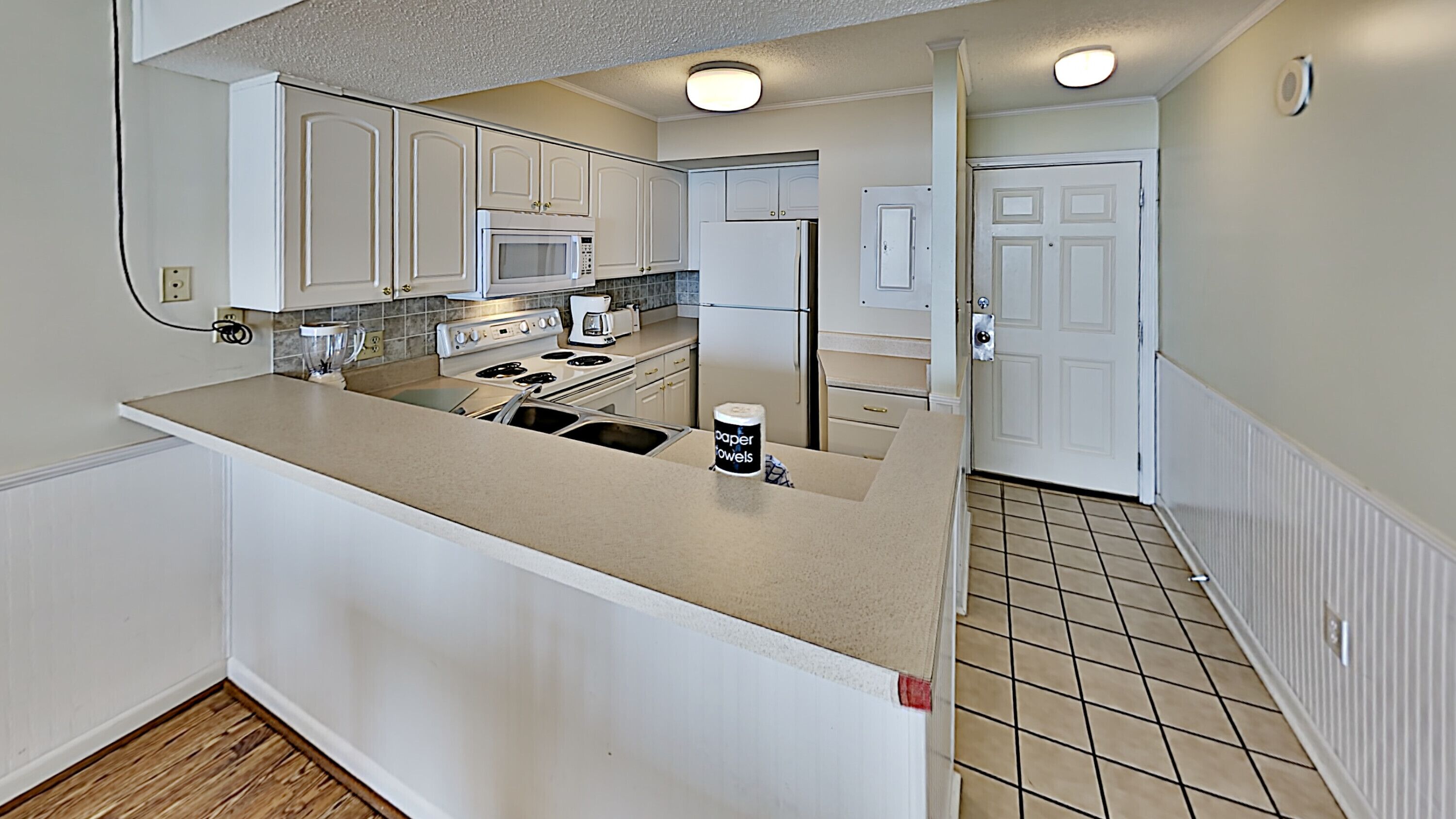 A kitchen with white cabinets, a stove, microwave, and refrigerator. There is a countertop with a paper towel dispenser.