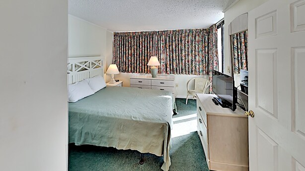 Quarto de hotel com cama, escrivaninha, cadeira, luminária e televisão.