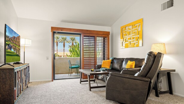 A living room with a leather sofa, a coffee table, a TV stand, and a door leading to a balcony with a view of palm trees. - Plaza Villas 615 (Palm Springs)