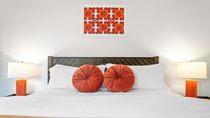 A neatly made bed with white linens, two red pillows, and a wooden headboard. There are two bedside lamps with orange bases. A colorful abstract artwork is displayed above the bed. - Plaza Villas 615 (Palm Springs)