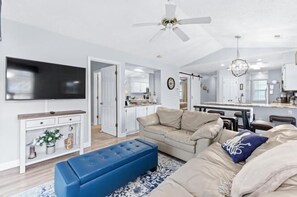 A modern living room with a beige sofa, blue ottoman, and a flat-screen TV mounted on the wall. - River Oaks 28G (Myrtle Beach)