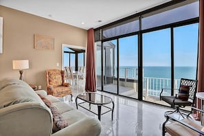 A spacious living room with a large window offering a view of the sea, a sofa, a chair, and a small table. - Sands Beach Club Ph1114 (Myrtle Beach)