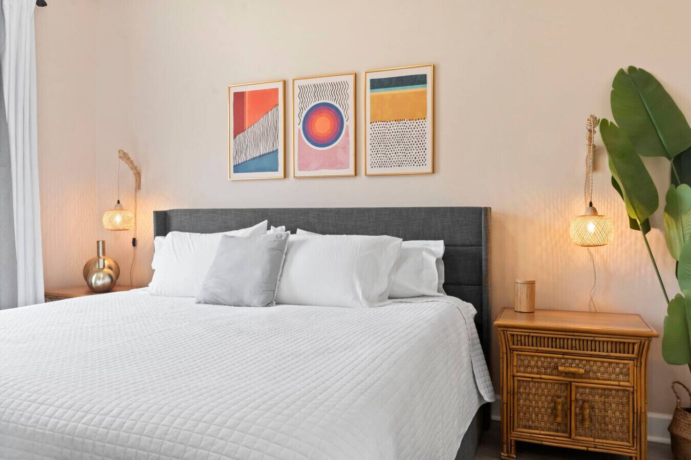Un lit bien fait, avec une literie blanche, une tête de lit rembourrée de couleur grise et une table de chevet sur laquelle se trouve un panier en osier. Trois œuvres d’art encadrées sont accrochées au mur au-dessus du lit.
