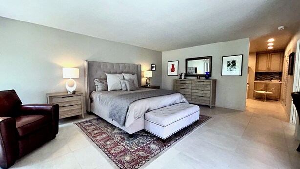 A bedroom with a large bed, bedside tables with lamps, a chair, a dresser, and a small kitchenette. - Villa Cha Cha (Rancho Mirage)