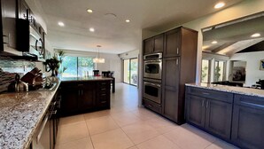 A modern kitchen with dark cabinetry, a central island, stainless steel appliances, and a large window providing natural light. - Villa Cha Cha (Rancho Mirage)