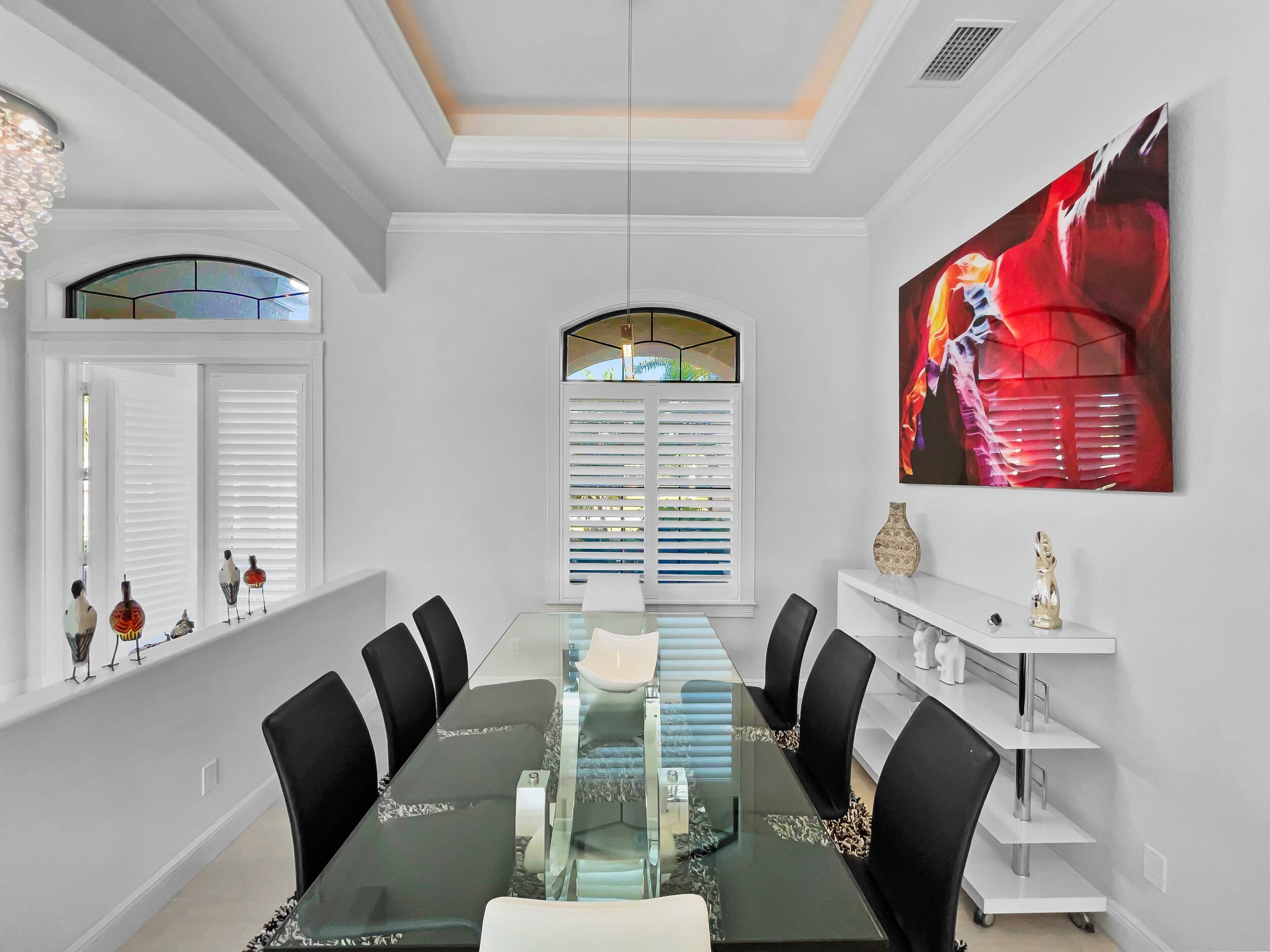 A dining area with a glass table, black chairs, white walls, a large painting, and a window with blinds.