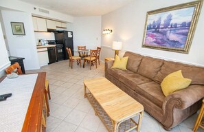 Ein Wohnzimmer mit braunem Sofa, Couchtisch aus Holz und Essbereich mit Stühlen. Im Hintergrund ist eine Küche mit Kühlschrank und Herd zu sehen.
