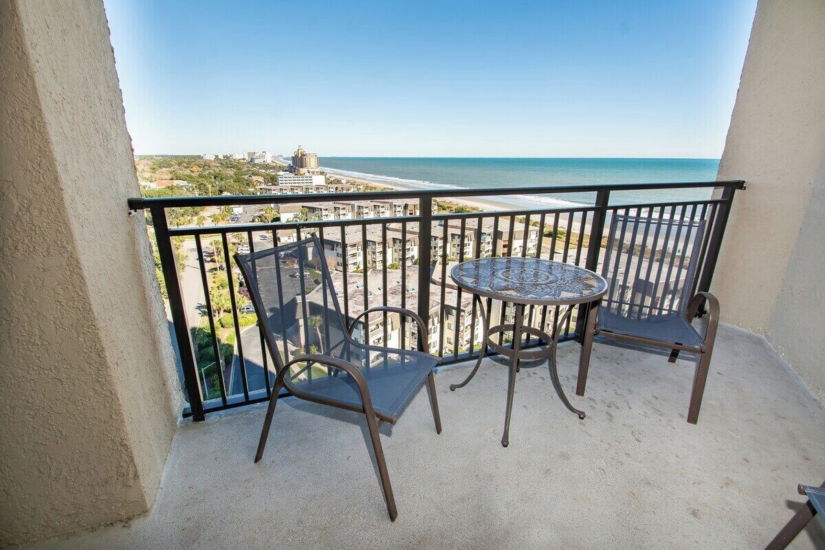 Een balkon met een tafel en stoelen, met uitzicht op een strand en stadsgezicht.