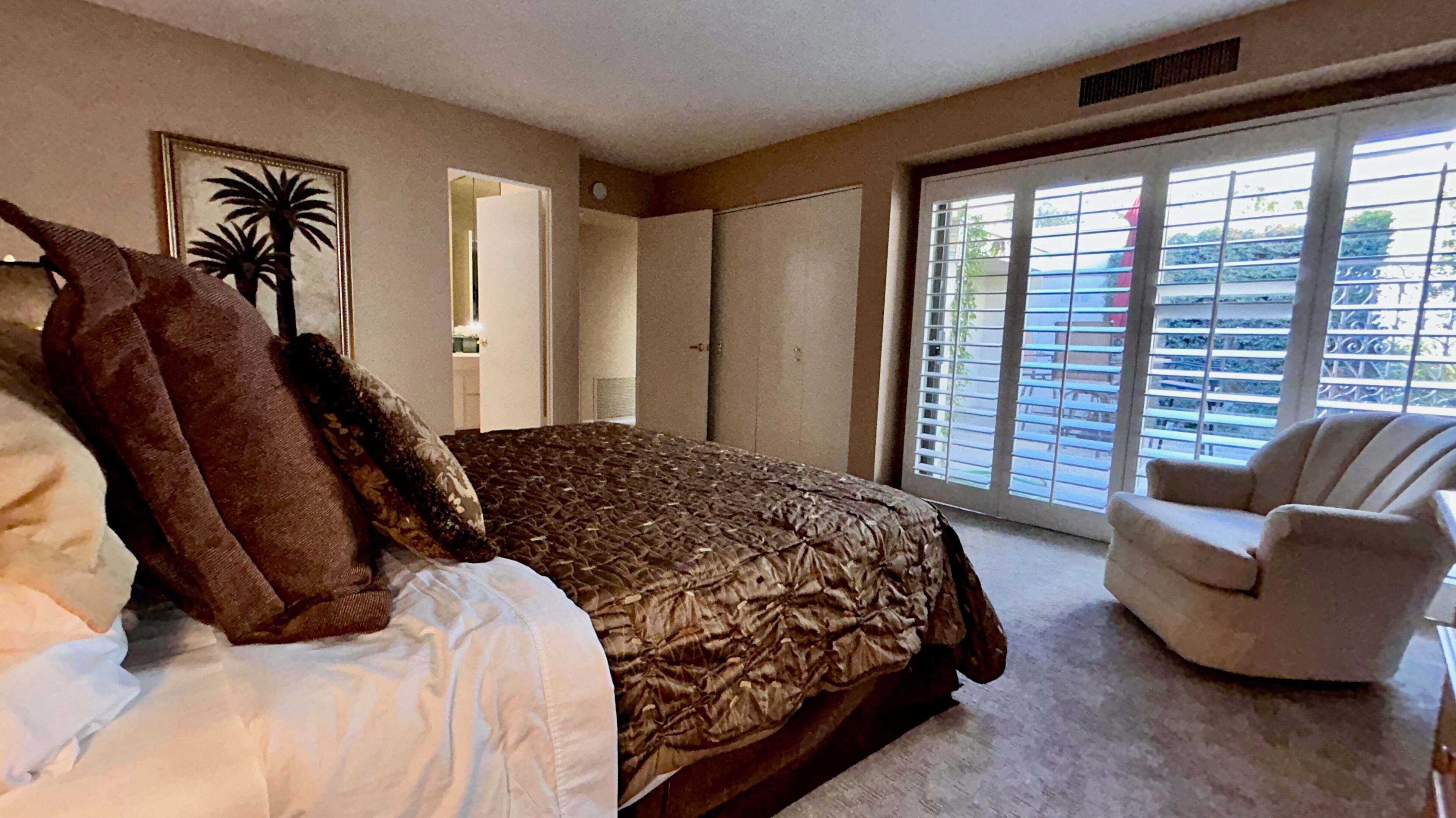 A bedroom with a bed, a chair, a large window with blinds, a door, and a framed picture on the wall.