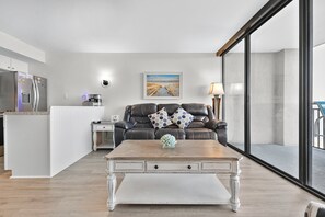 A modern living room with a wooden coffee table, a black leather sofa with patterned cushions, a white side table, and a painting on the wall. - Ocean Forest Plaza Unit 1412 (Myrtle Beach)