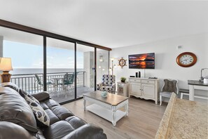 A modern living room with a large glass door leading to a balcony, a sofa, a coffee table, and a view of the ocean. - Ocean Forest Plaza Unit 1412 (Myrtle Beach)