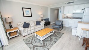 A living room with a sofa, coffee table, and a kitchen area with cabinets and appliances. - Ocean Forest 1006 (Myrtle Beach)