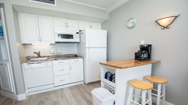 A compact kitchen with white cabinets, a microwave, a refrigerator, a coffee maker, and two stools. - Ocean Forest 1006 (Myrtle Beach)