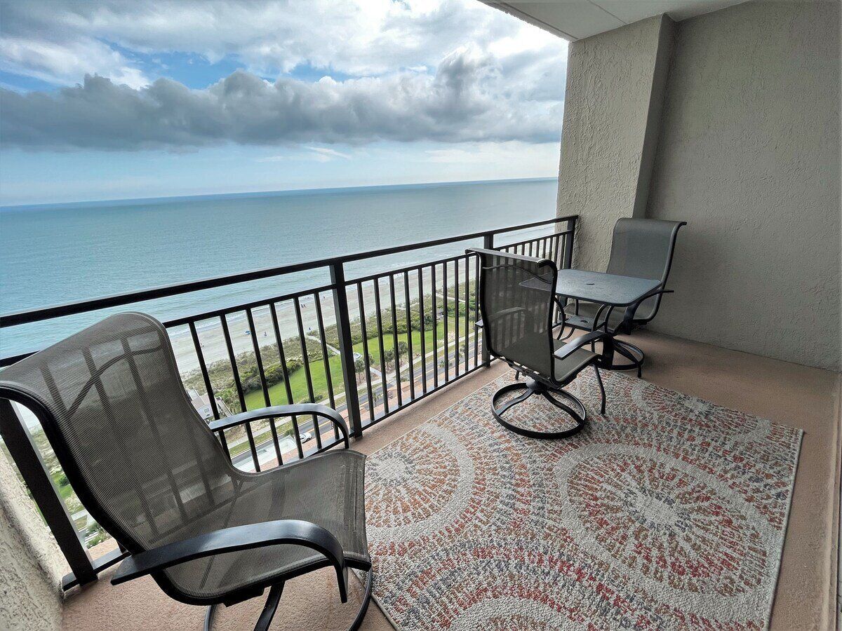 A balcony with a view of the sea, two chairs, a table, and a patterned rug.