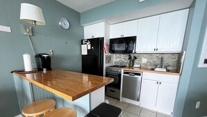 Ett kompakt kök med en liten bardisk med trästolar, ett kylskåp, en mikrovågsugn, en spis och en diskmaskin.