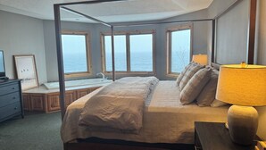 A four-poster bed with a view of the sea, a ceiling fan, and a bedside lamp.