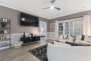A modern living room with a flat-screen TV, a ceiling fan, and a large window with curtains. - Oh My Gosh So Posh! Stylish 2BR Condo w/ Glam Decor, Bright Spaces & Relaxed Comfort (Myrtle Beach)