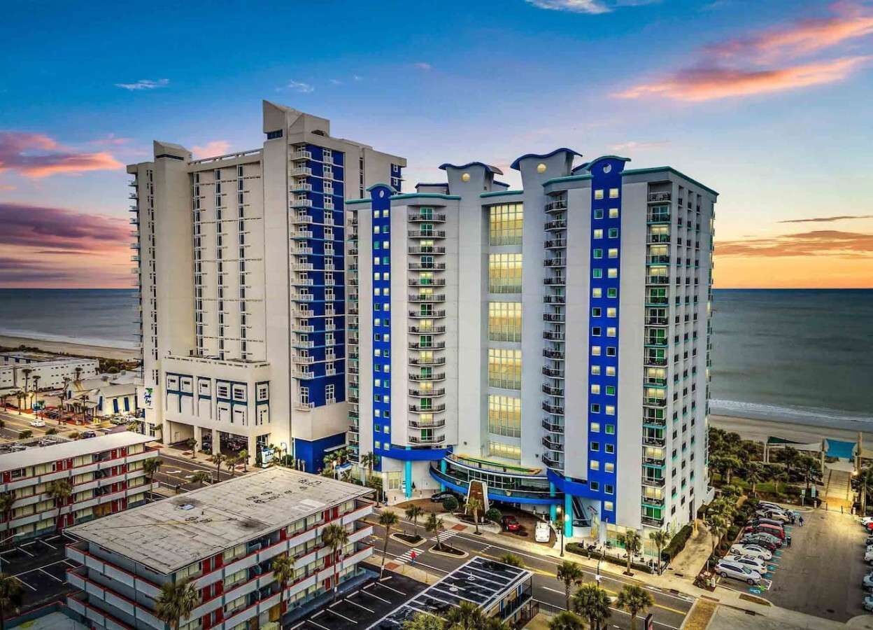 A high-rise hotel with multiple floors, a parking area, and a view of the beach.