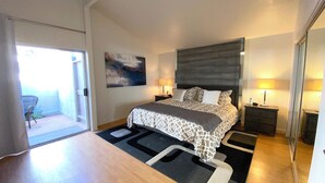 Une chambre à coucher comprenant un lit, deux tables de chevet, une commode, une chaise, un tableau et une porte-fenêtre coulissante donnant sur un balcon.