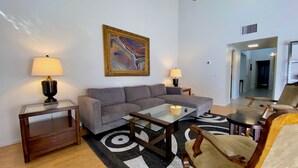 Un salon moderne avec un canapé gris, une table basse en verre et un tapis à motifs. On y trouve deux lampes sur des tables d’appoint et un tableau au mur.