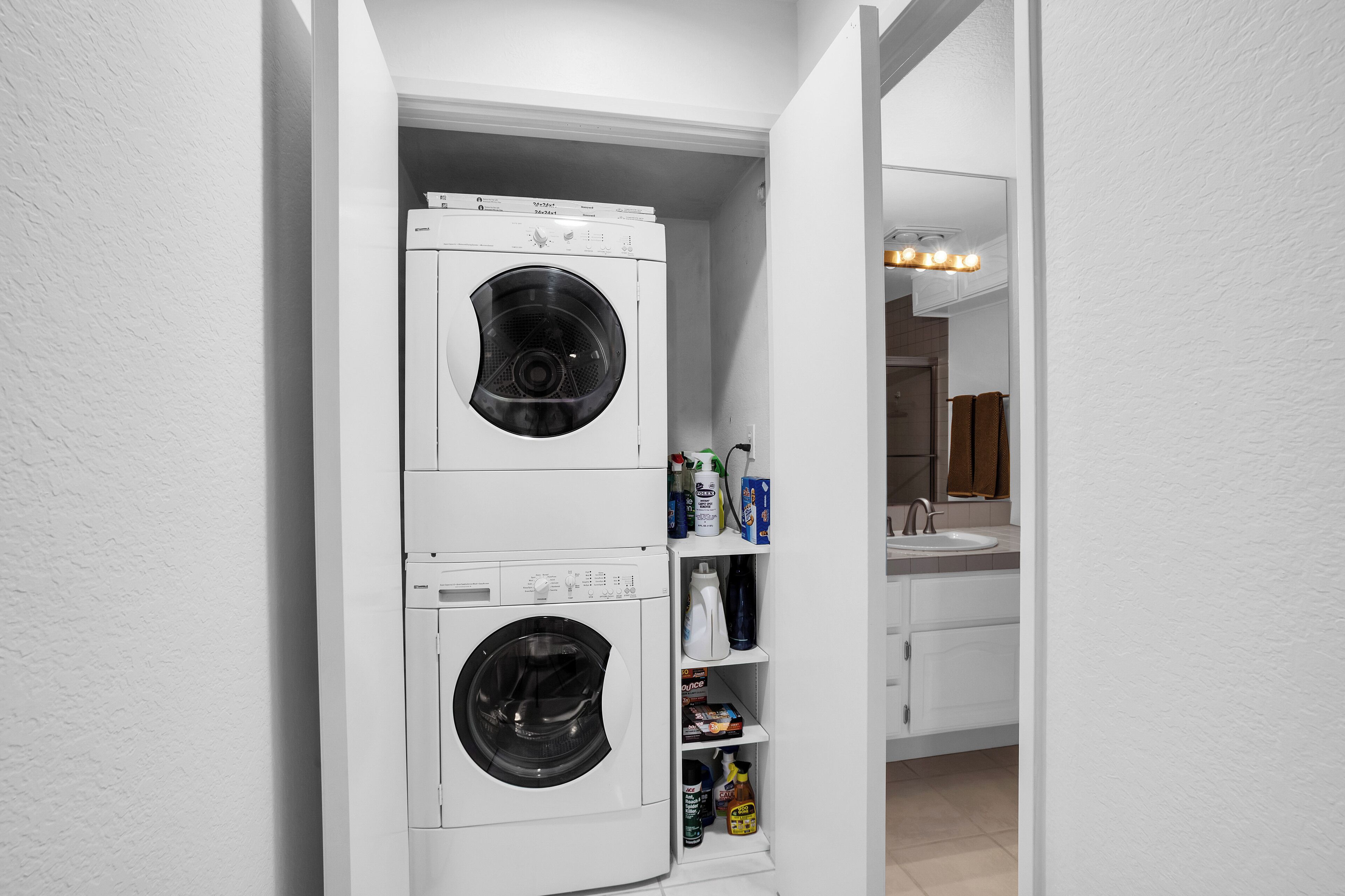 Un espace buanderie compact comprenant une machine à laver et un sèche-linge superposés, une étagère avec des produits de nettoyage et une salle de bain avec un lavabo et un miroir.
