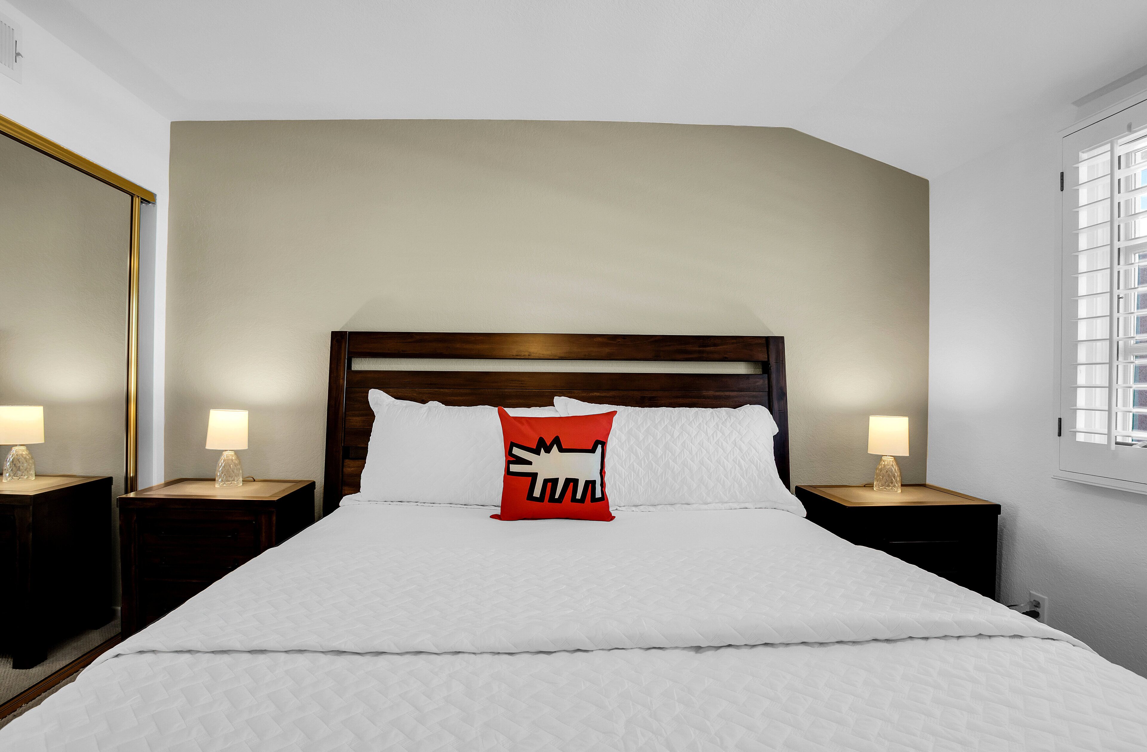 A bedroom with a large bed, two bedside tables with lamps, a wooden headboard, and a window with blinds.