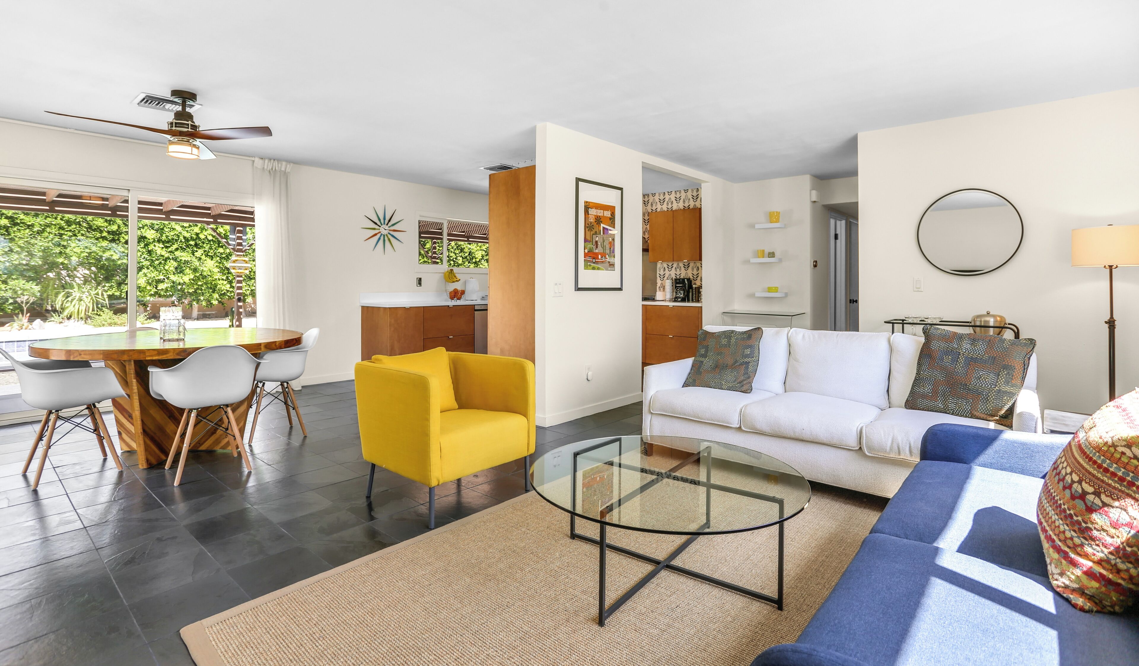 A modern living room with a yellow armchair, a glass coffee table, a white sofa, and a blue sofa. There is a dining area with a wooden table and chairs, and a kitchen in the background.