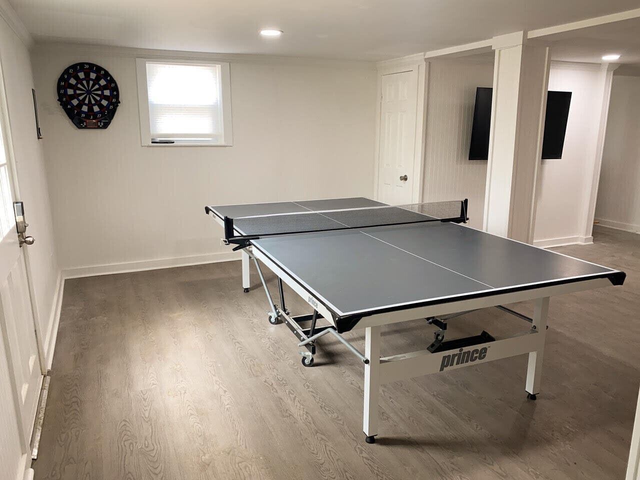 Une table de ping-pong dans une pièce où se trouvent un jeu de fléchettes, une fenêtre et une porte.