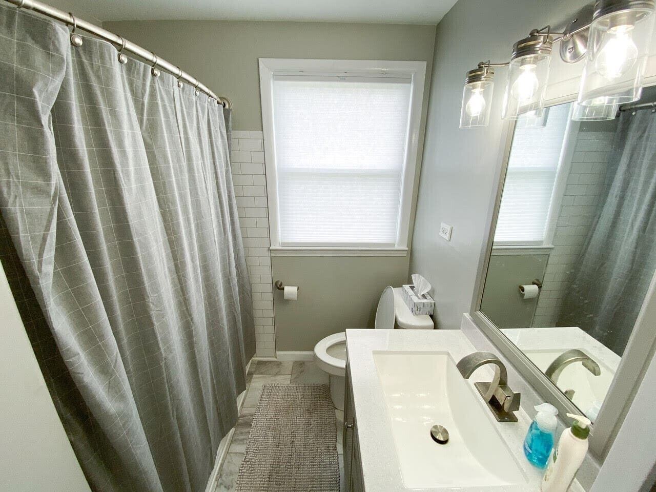 Une salle de bain avec un rideau de douche, des toilettes, un lavabo avec un miroir et une fenêtre avec des stores.