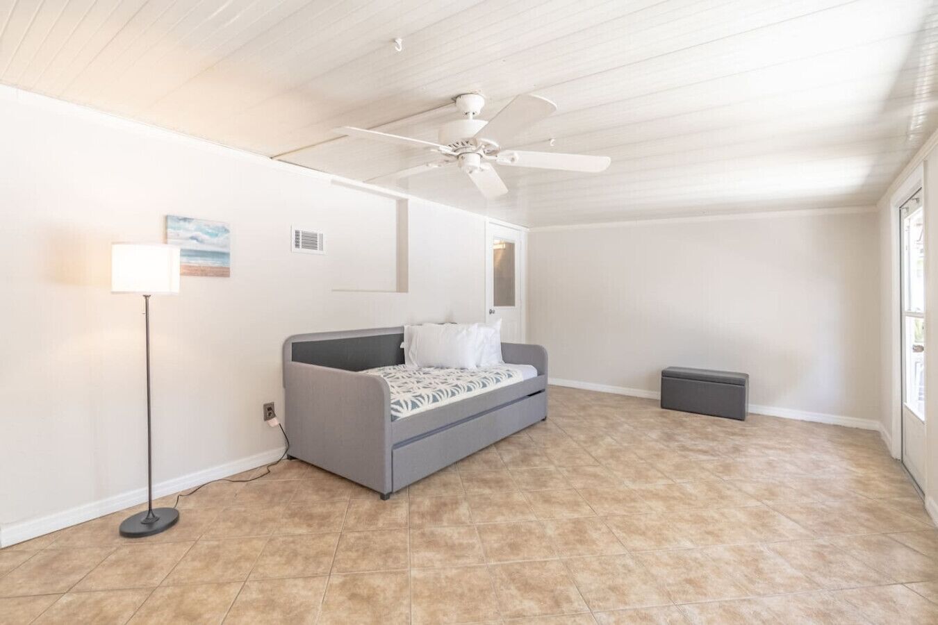 A spacious room with a gray sofa, a standing floor lamp, a ceiling fan, and a large window.