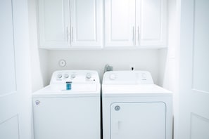 A laundry room with two white front-loading washers and dryers, white cabinets above, and a small window. - Beautiful, Pool, Gym, Near Theheights, BBQ (Houston)